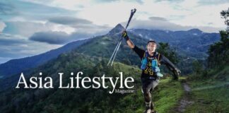 A trail runner raises his poles in celebration on a ridge path, forested mountains behind