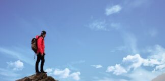 A hiker in a red jacket stands at the edge of a rocky outcrop, looking out over cloud and distant mountains