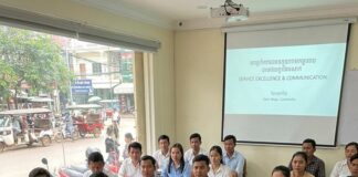 A trainer addresses a classroom of Cambodian hospitality students beside a Service Excellence and Communication slide