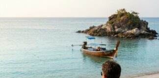 Phuket Expat’s Honest Lifestyle Audit vs Dubai: A Comprehensive Comparison of Living Standards and Experiences A man sits alone at a wooden beach table with coffee, looking out at a longtail boat and a small rocky island