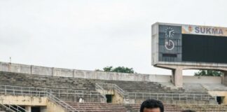 An official briefs a colleague with a clipboard beside an athletics track, a Sukma scoreboard behind