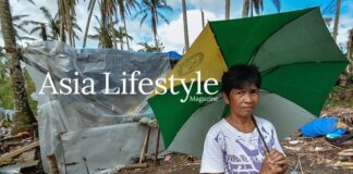 A woman holds a green umbrella beside a tarpaulin shelter amid storm debris and stripped palm trees